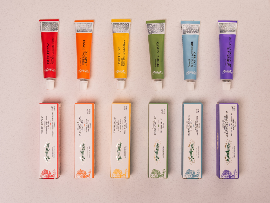 Six tubes of body butter and their matching boxes arranged in two rows on a light pink background. The tubes are red, orange, yellow, green, light blue, and purple, with white labels.
